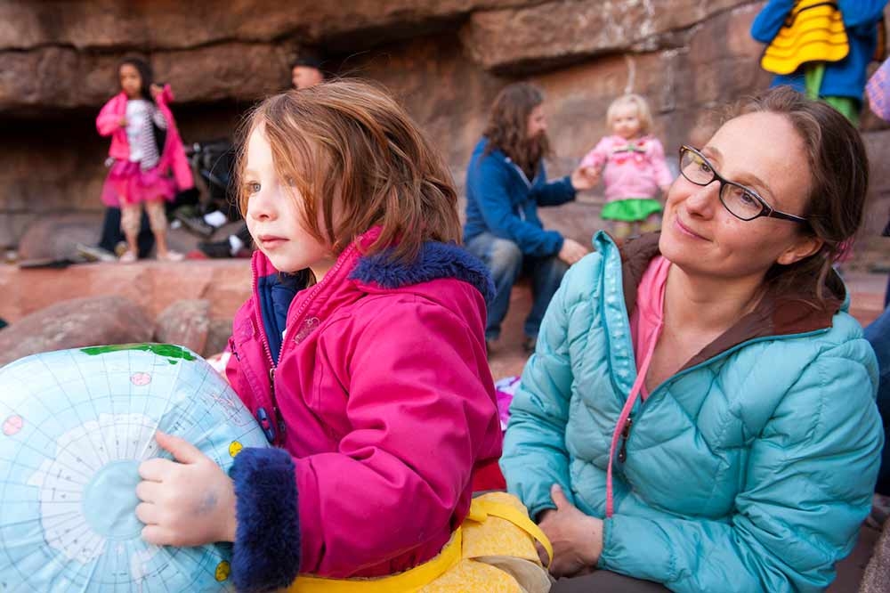 Adult in a blue coat and child in a pink coat listening to a story outside. Child is holding a toy globe.