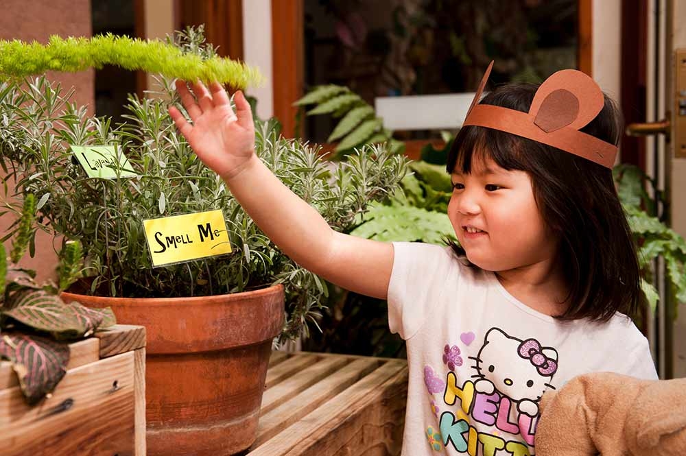 Child wearing brown paper headband with cut out animal ears. They are touching a plant. The potted plant has a sign stuck into the dirt that says 'smell me'.
