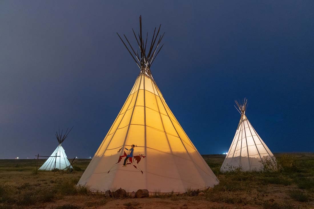 Three tipis on the plains with a dark sky behind them. 