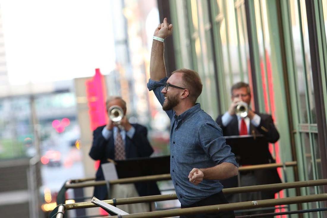 Man with arm raised and two men with trumpets in background