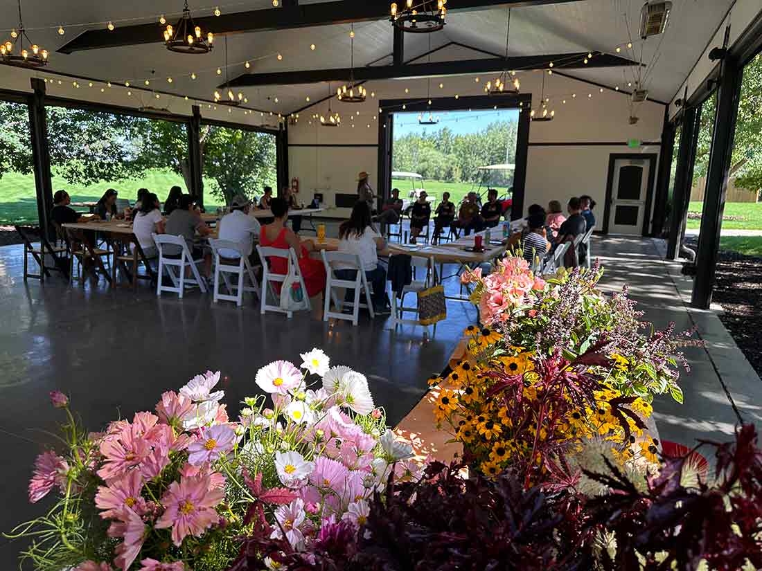 People sitting inside a building, listening to a speaker. In the foreground are colorful flower arrangements. 