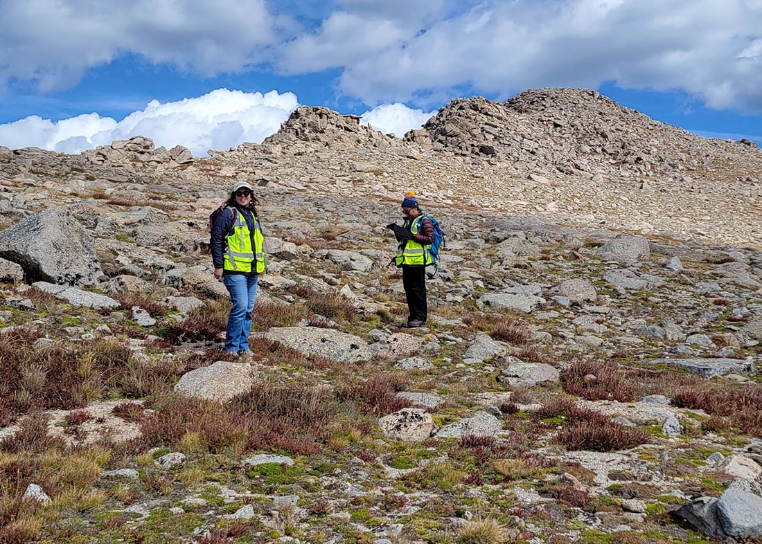 Two people on a rocky mountain slope with mountain peaks and clouds in background