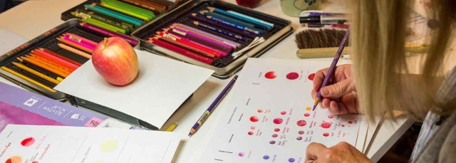 Person's hand holding a purple colored pencil. In the background are an apple and a set of colored pencils.