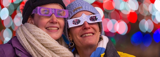 Two smiling people wearing Holospex glasses, hats and scarves, are looking at holiday lights. Blurred colorful lights are behind them.