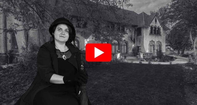 Woman wearing black clothes and black gloves, holding a closed black umbrella, in the foreground; mansion in the background at night